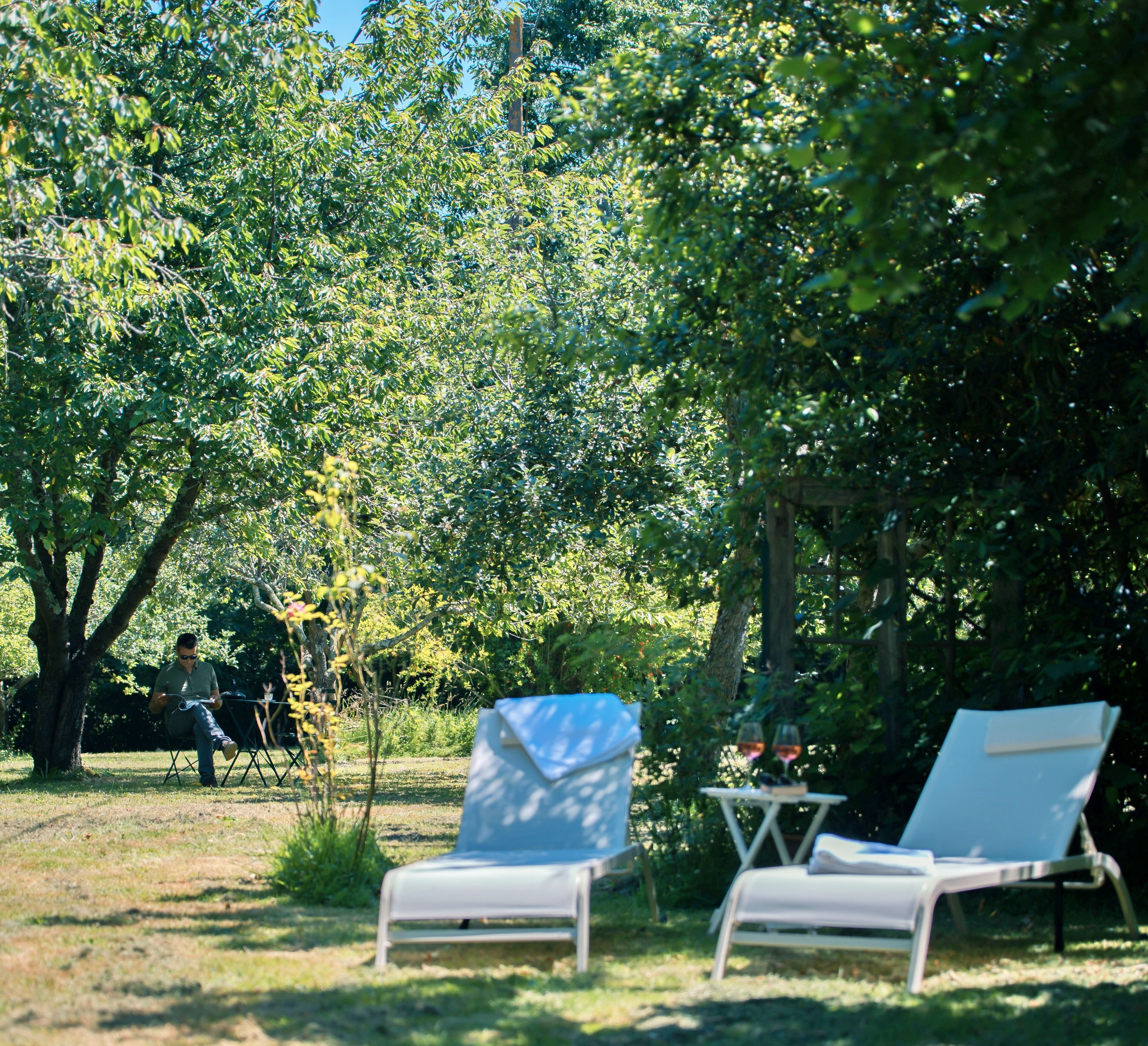 A serene outdoor seating area with two lounge chairs, one with a white cover and another without, set against a backdrop of lush green trees and a clear sky.