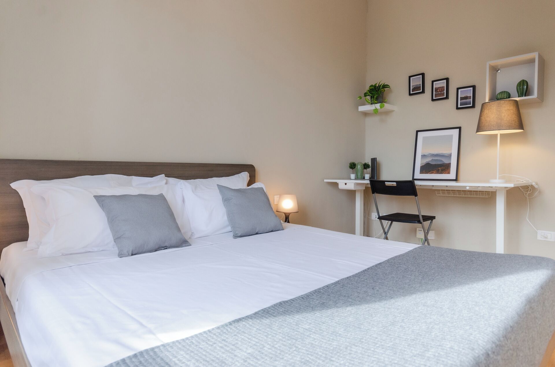 A neatly made bed with white and gray linens, a bedside lamp, a desk with a chair, and a small shelf with plants and pictures.