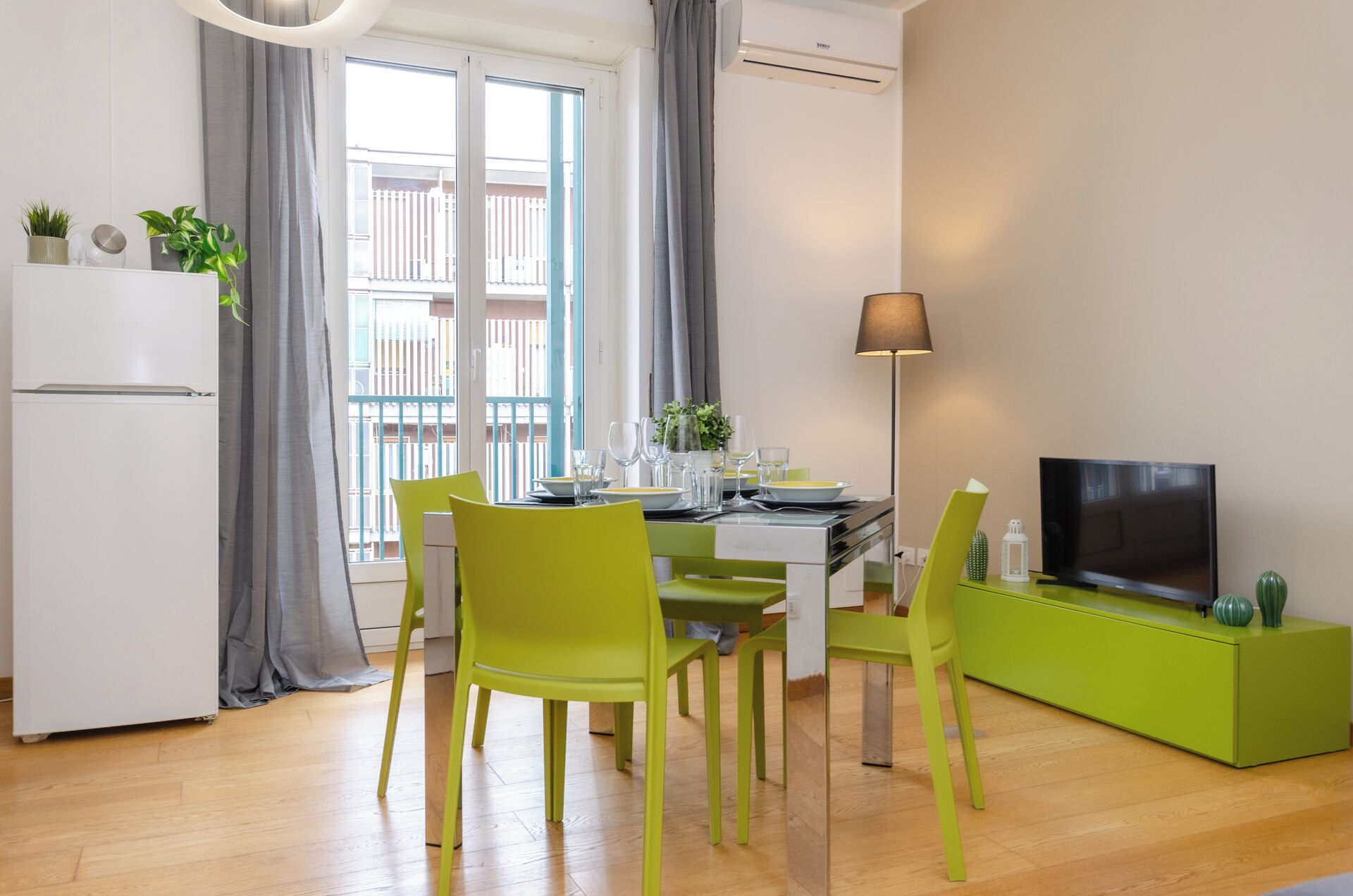 A modern dining area with a table set for four, green chairs, a white refrigerator, a flat-screen TV on a green stand, and a window with grey curtains.