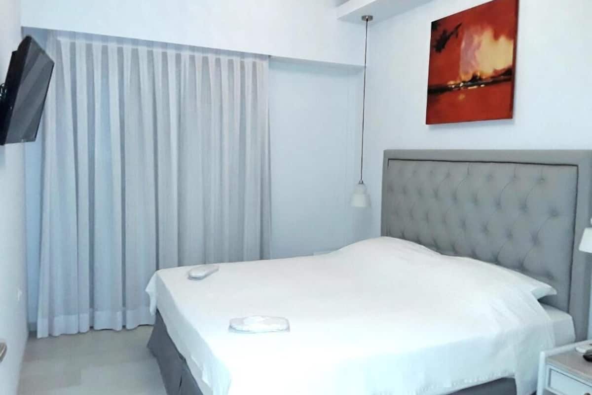 A hotel room with a bed, a flat-screen TV mounted on the wall, a painting above the bed, and a window with curtains.