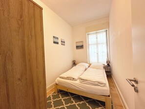 A small, narrow room with a wooden door, a bed with white bedding, a small table with a lamp, and a window with sheer curtains.