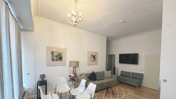 Una sala moderna con una mesa de comedor de vidrio, sillas blancas, un sofá gris, una araña y obras de arte en las paredes.