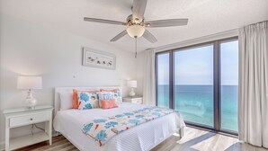 A bedroom with a large bed, a ceiling fan, a nightstand with a lamp, a window with a view of the ocean, and a framed picture on the wall. - Unbeatable Views in this 3BR 3BA Condo at the Watercrest w/Beach Chairs! (Panama City Beach)