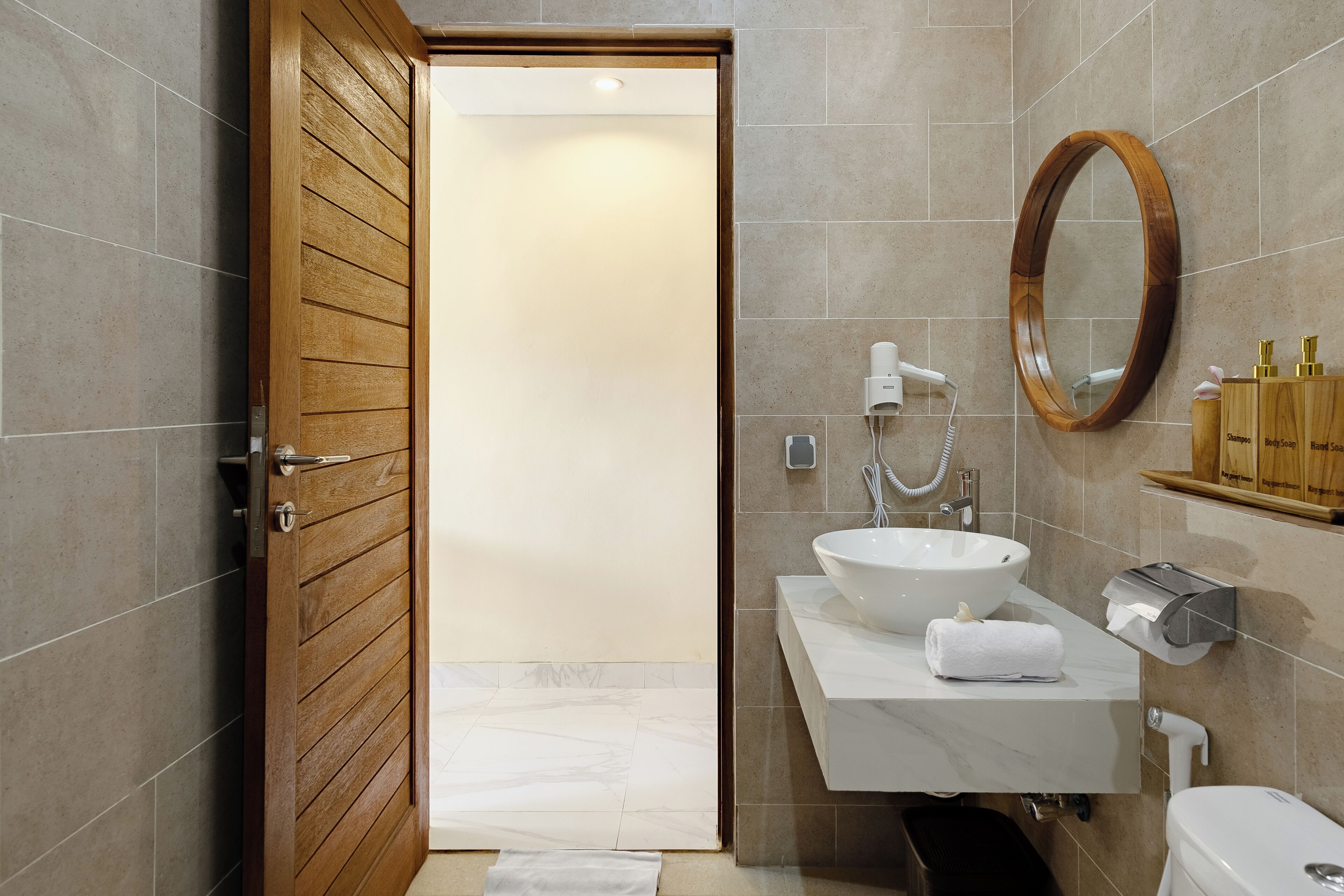 Une salle de bain moderne avec une porte en bois, un lavabo blanc, un miroir et des articles de toilette rangés sur une étagère.