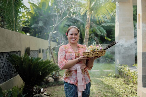 A person holding a tray with incense sticks and flowers, standing in a garden with greenery and a stone wall. - Ray Guest House (Ubud)