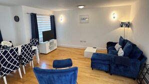 Una sala de estar moderna con televisor, un sofá azul, una mesa de comedor con sillas estampadas y una pequeña mesita auxiliar blanca.