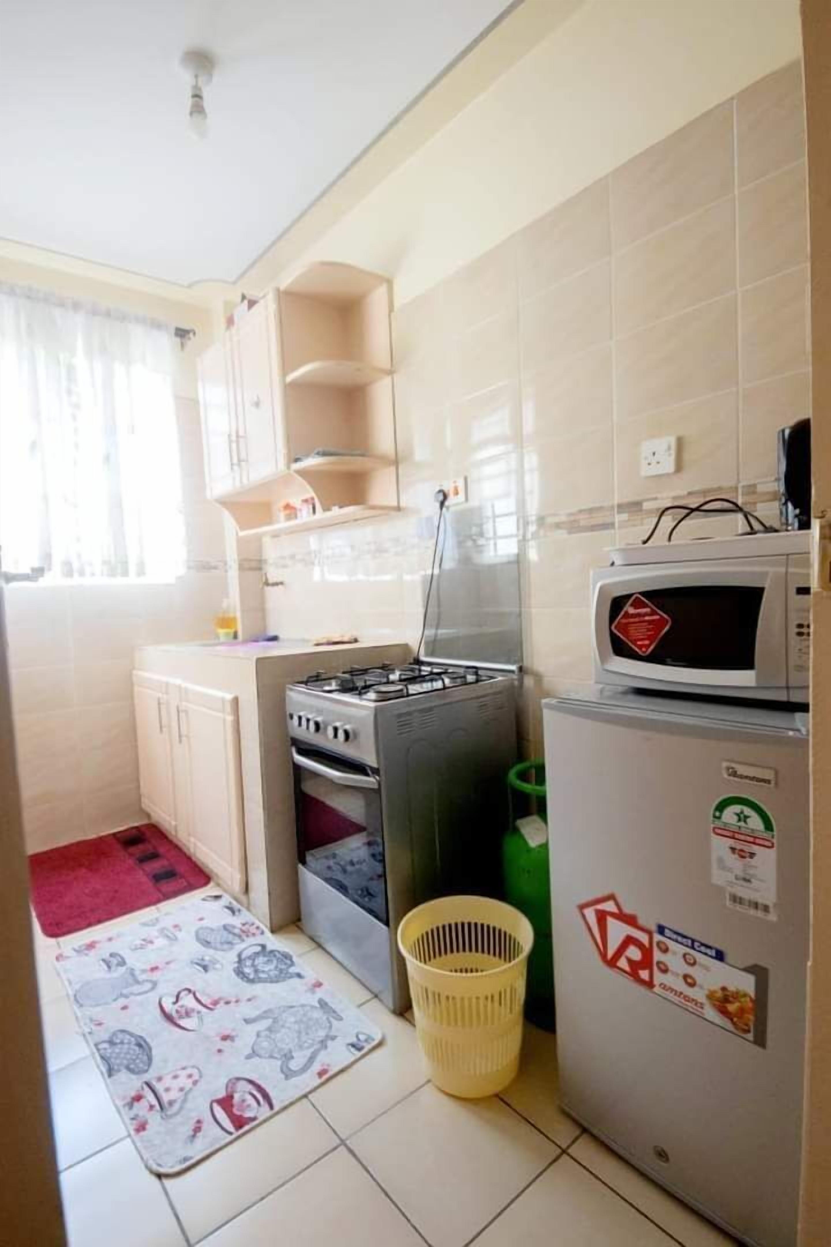 A compact kitchen with a gas stove, microwave, and refrigerator. There is a small window with curtains, a red rug, and a basket on the floor.
