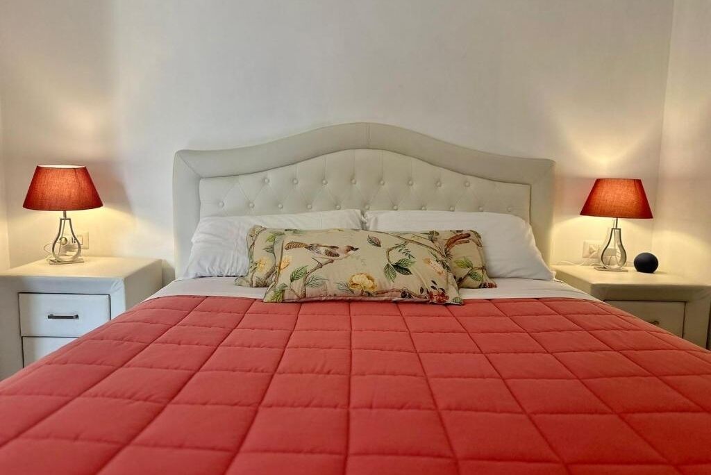 A neatly made bed with a floral pillow, a red quilt, and two bedside lamps on white nightstands.