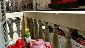 Una calle estrecha flanqueada por edificios, un letrero que dice 'IL SISTINA', y una mesa con una tetera, tazas y un cactus.