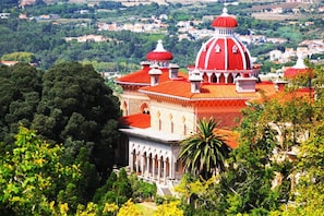 A large building with a prominent red dome and multiple smaller domes, surrounded by lush greenery and a hilly landscape.
