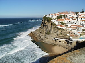 A coastal town with white buildings and red roofs, a rocky shoreline, and waves crashing onto the beach.