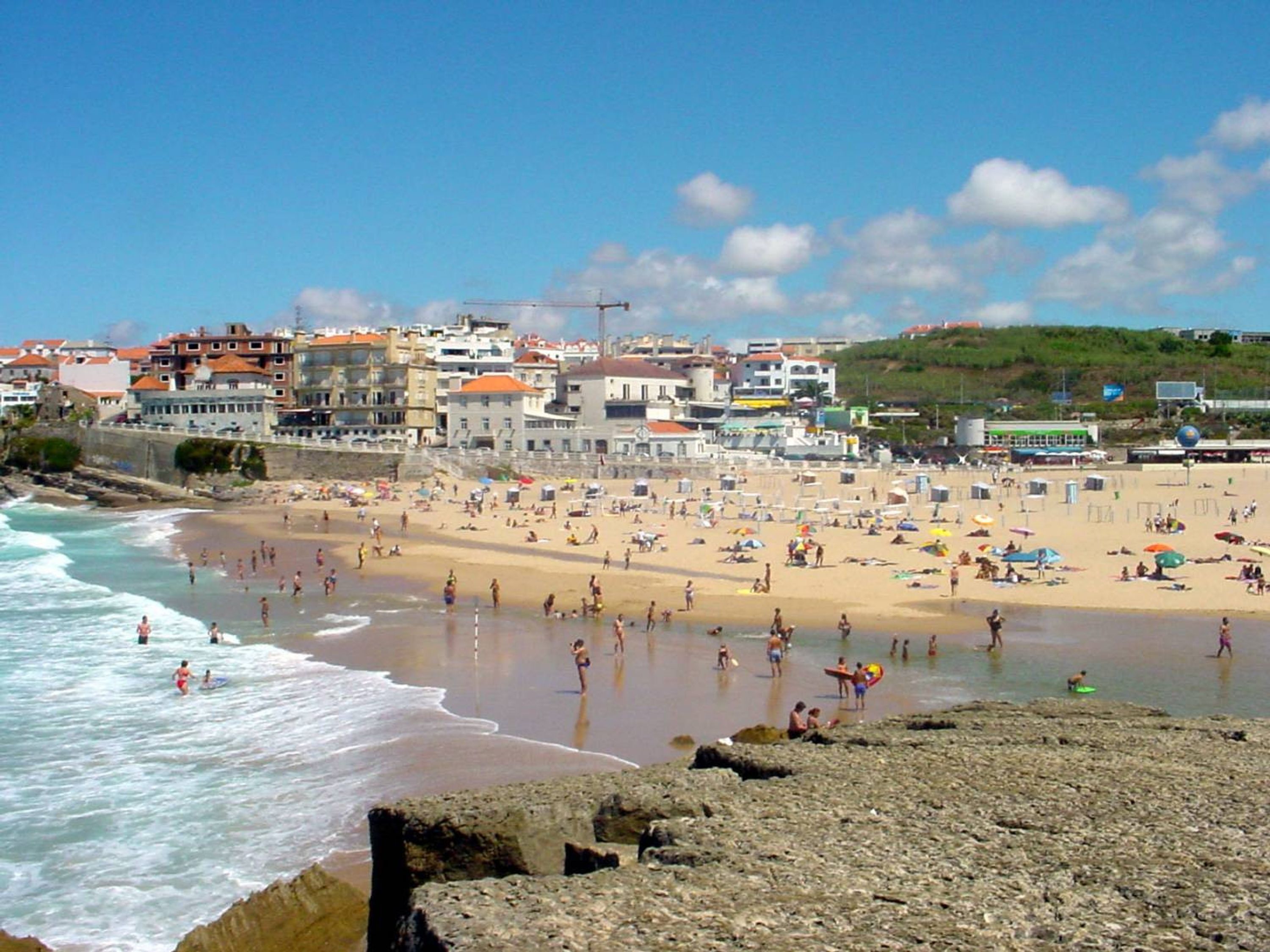 Una playa con gente nadando y tomando el sol, edificios al fondo y una costa rocosa.