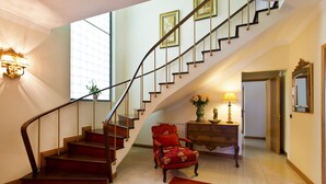 A grand staircase with a wooden handrail, a red upholstered chair, and a lamp on a wooden side table.