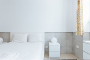 Ett modernt, minimalistiskt sovrum med en stor säng, ett sängbord med en lampa och ett litet bord.
