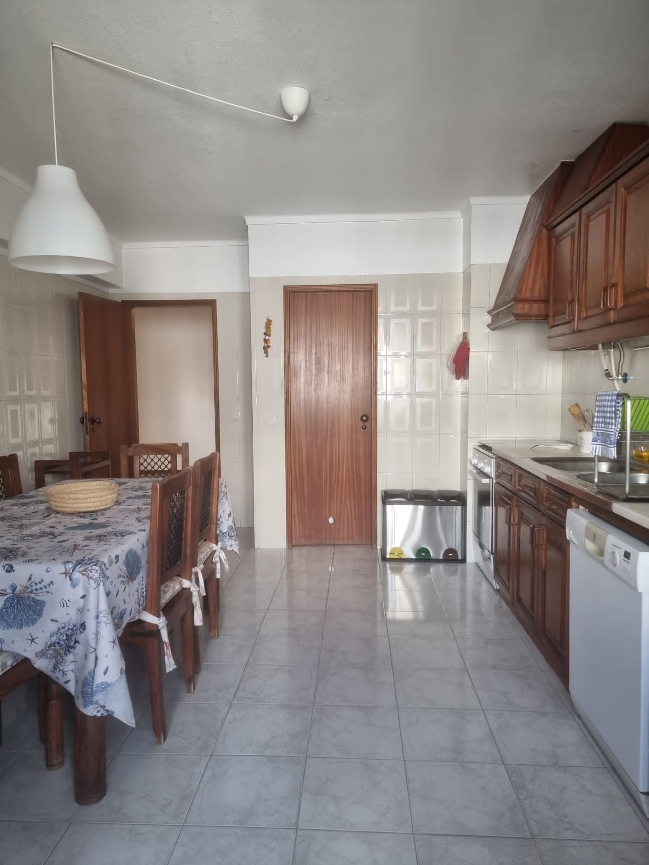 A kitchen with wooden cabinets, a dining table with chairs, a hanging light, and a trash bin.
