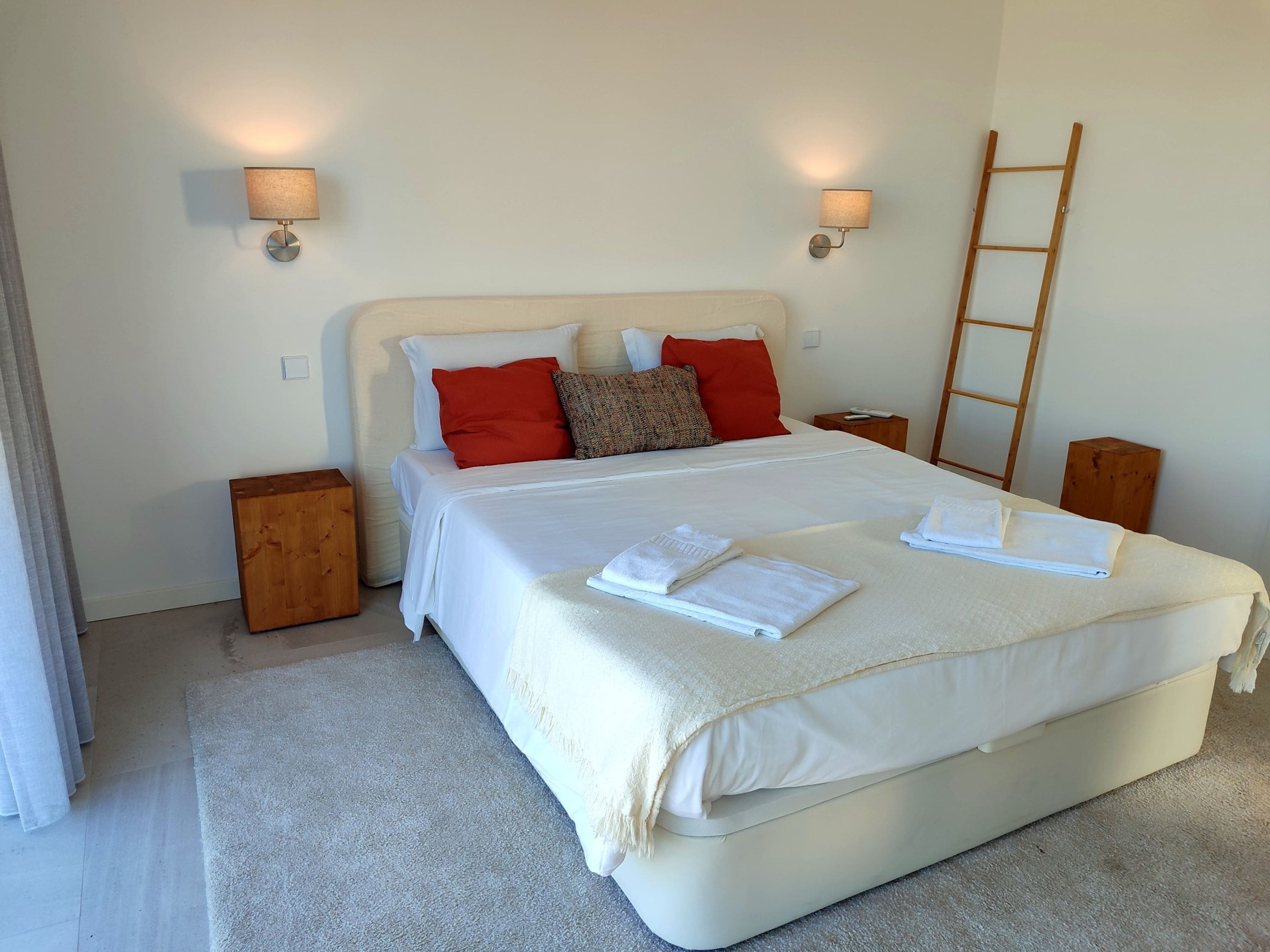 Cama bem arrumada com lençóis brancos e almofadas vermelhas, duas mesas de cabeceira com abajures, uma escada de madeira e piso coberto por carpete.