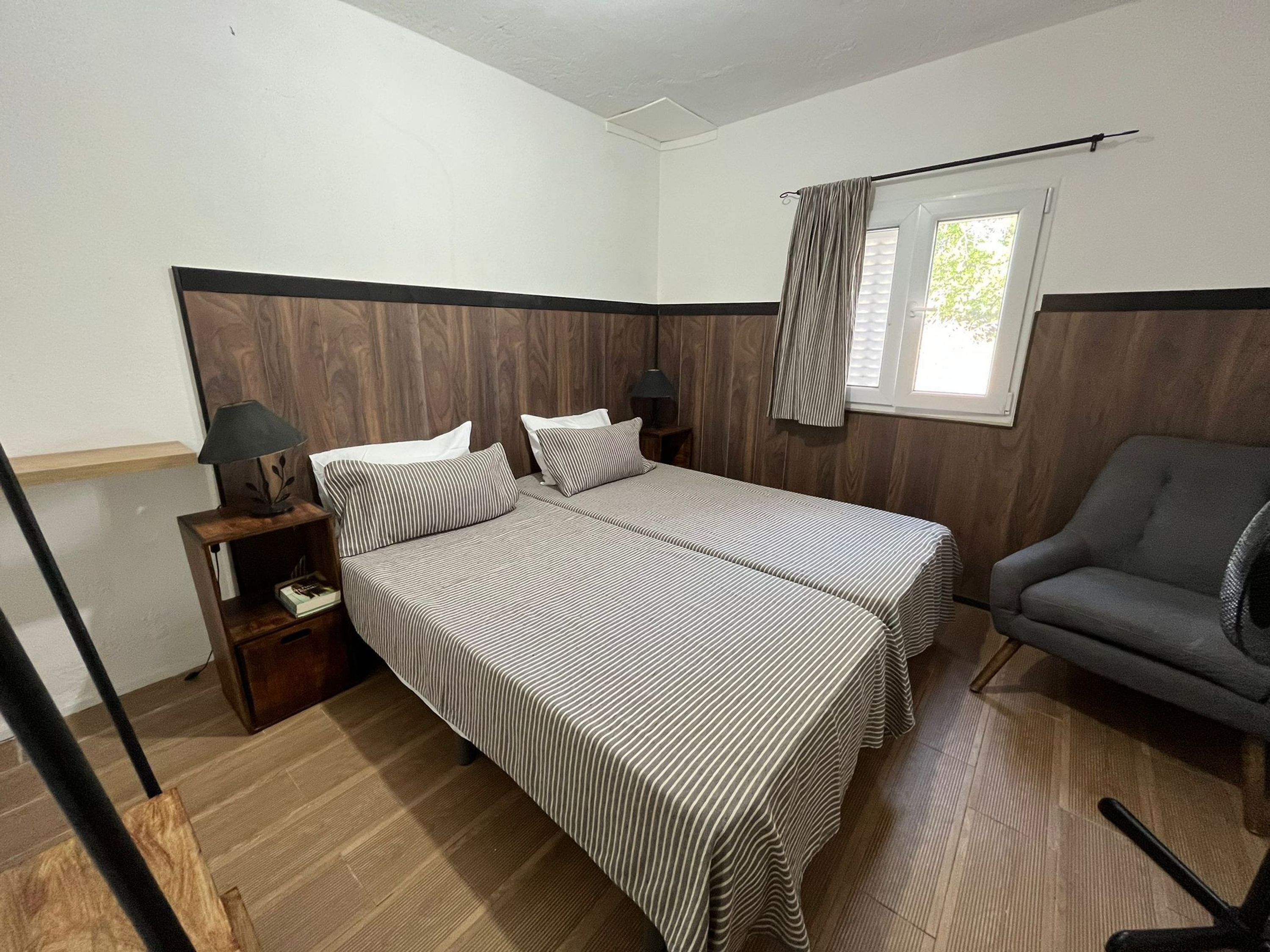 Une chambre avec deux lits, une chaise, une table de chevet avec une lampe et une fenêtre avec des rideaux.