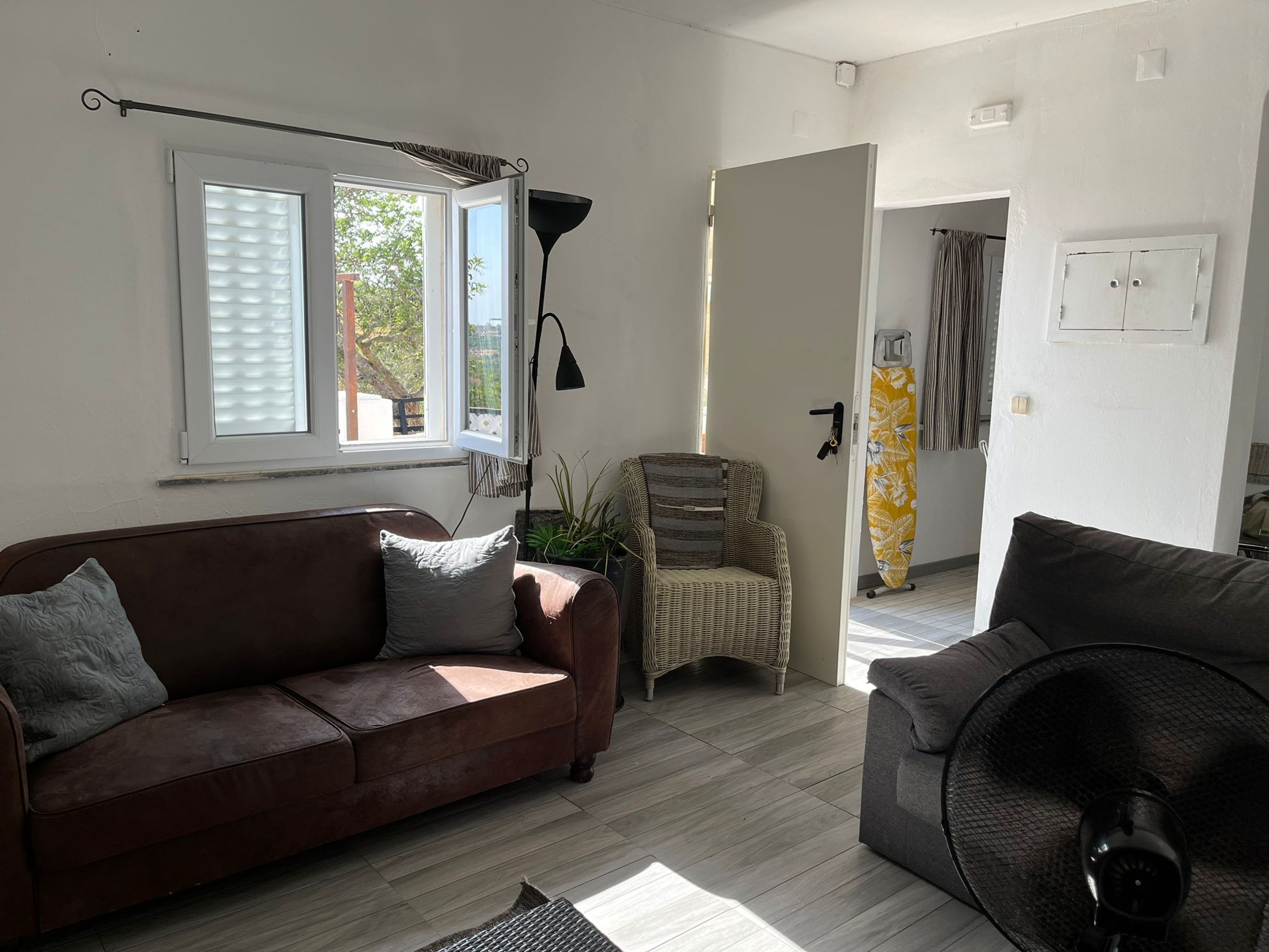 A living room with a brown sofa, a wicker chair, a fan, and a window with blinds.