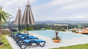 A poolside area with lounge chairs and umbrellas, a hanging chair, and a view of a town and hills in the background.
