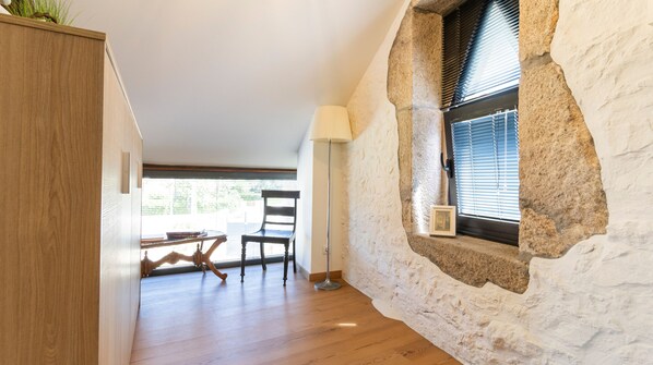 A room with a stone wall, a wooden floor, a table with chairs, a lamp, and a window with blinds.