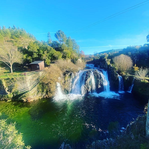 Una cascada que se precipita sobre rocas hacia un río, rodeada de árboles y un pequeño edificio.
