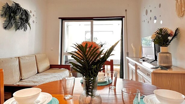 A dining area with a wooden table set for four, a glass vase with a plant, and a hanging light fixture. - Nice Flat 150 mt Beach With Garage (Portimão)