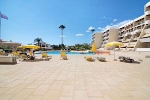 A poolside area with sun loungers and umbrellas, a clear sky, and a modern building with balconies.