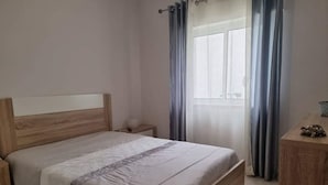 A bedroom with a wooden bed, a nightstand, a small table with a decorative item, and a window with curtains. - Ground Floor Palmeira Gal Villa With Pool (Albufeira)