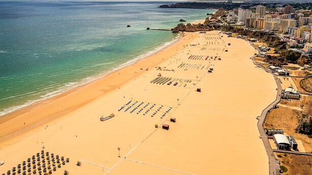 An aerial view of a beach with rows of sun loungers and umbrellas, a pier, and buildings in the background. - Superior Garage Balcony Praia da Rocha Prime Flat (Portimão)