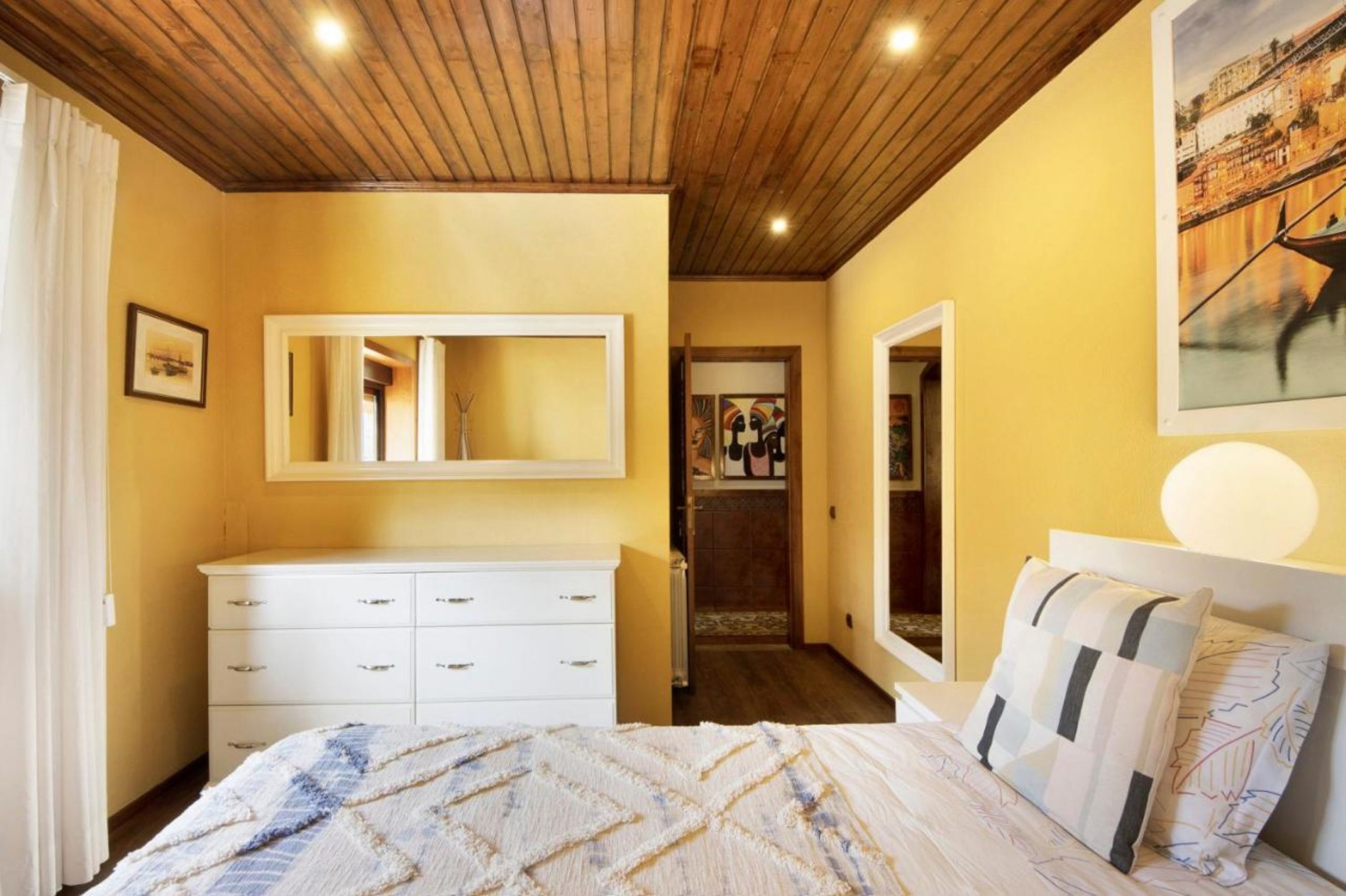 A bedroom with a wooden ceiling, a white dresser, a bed with patterned bedding, and a painting on the wall.