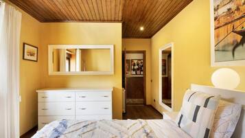 A bedroom with a wooden ceiling, a white dresser, a bed with patterned bedding, and a painting on the wall.