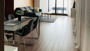 Un soggiorno moderno con zona pranzo, pavimento in legno, un divano, un tavolino da caffè e un'ampia finestra con tende.
