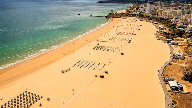 Una playa con filas de tumbonas y sombrillas, un paseo marítimo y edificios al fondo.