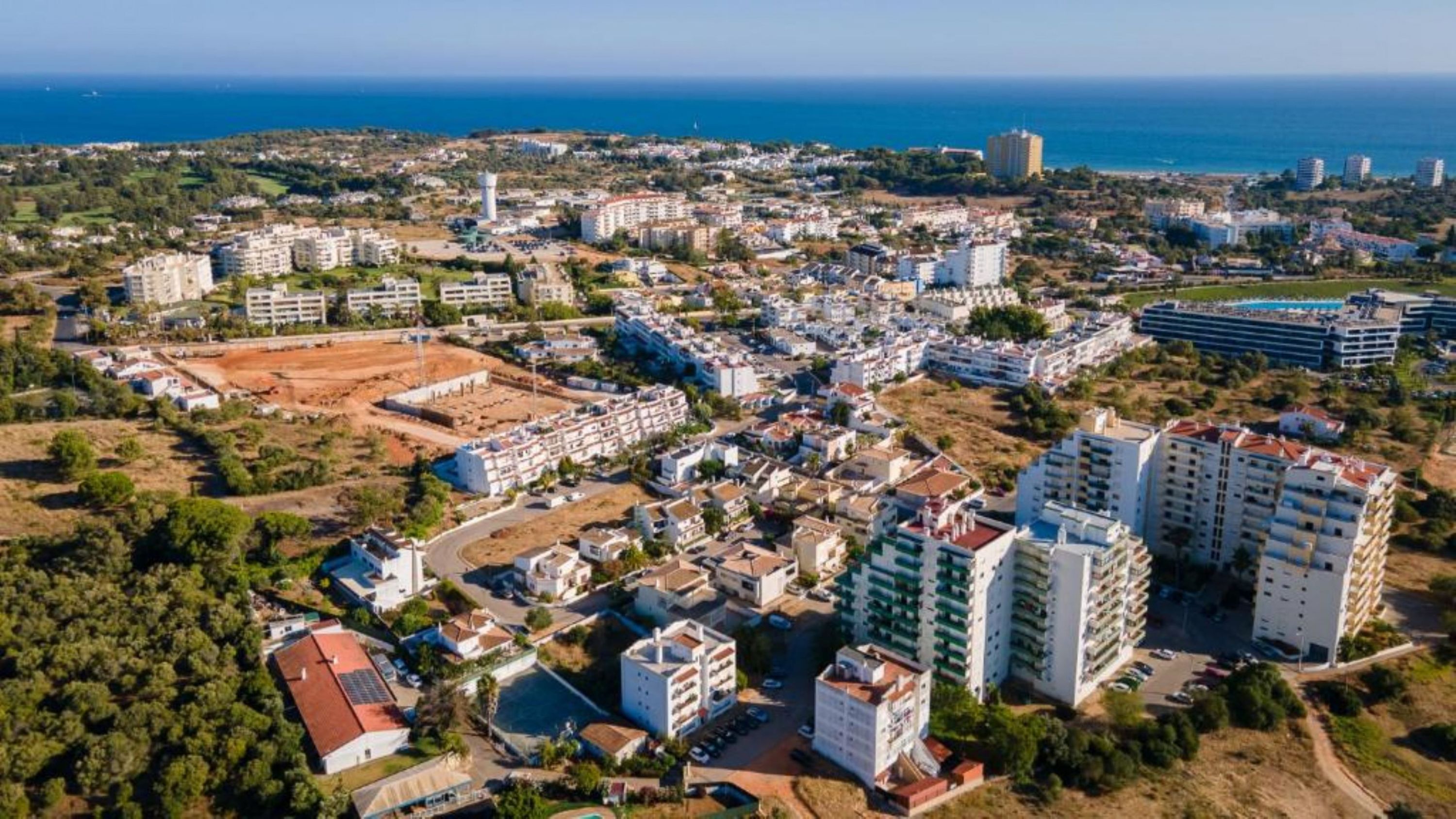 Vista aérea de un pueblo costero con edificios residenciales, un extenso campo abierto y una masa de agua al fondo.