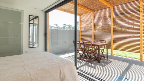 Ein Schlafzimmer mit einem Bett, einem großen Fenster und einer Glastür, die auf eine Terrasse mit Tisch und Stühlen führt.