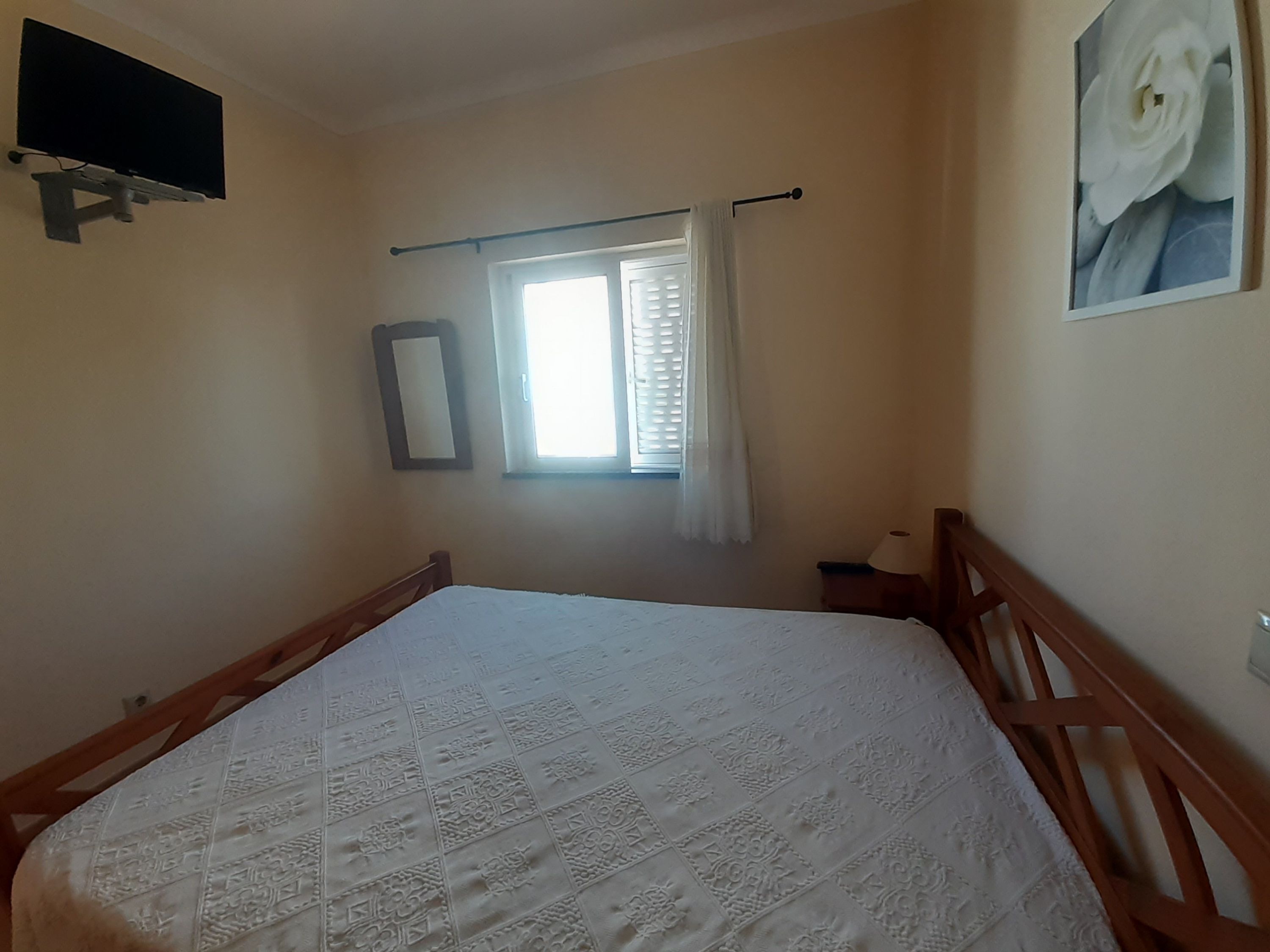 Ein Schlafzimmer mit einem Bett, einem Fenster mit Vorhängen, einem an der Wand montierten Fernseher und einem gerahmten Bild.