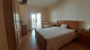 Un dormitorio con una cama grande, una mesita de noche de madera, un escritorio y una silla. Hay una puerta corrediza de vidrio que da a un balcón.