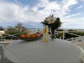 Ein Tisch mit Obstschale, zwei Gläsern mit Getränken und einem Blumenstrauß auf einer Terrasse mit Aussicht.