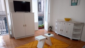 Una sala de estar con un televisor sobre un mueble bajo, una mesita blanca, un tapete amarillo y un aparador blanco con estantes.