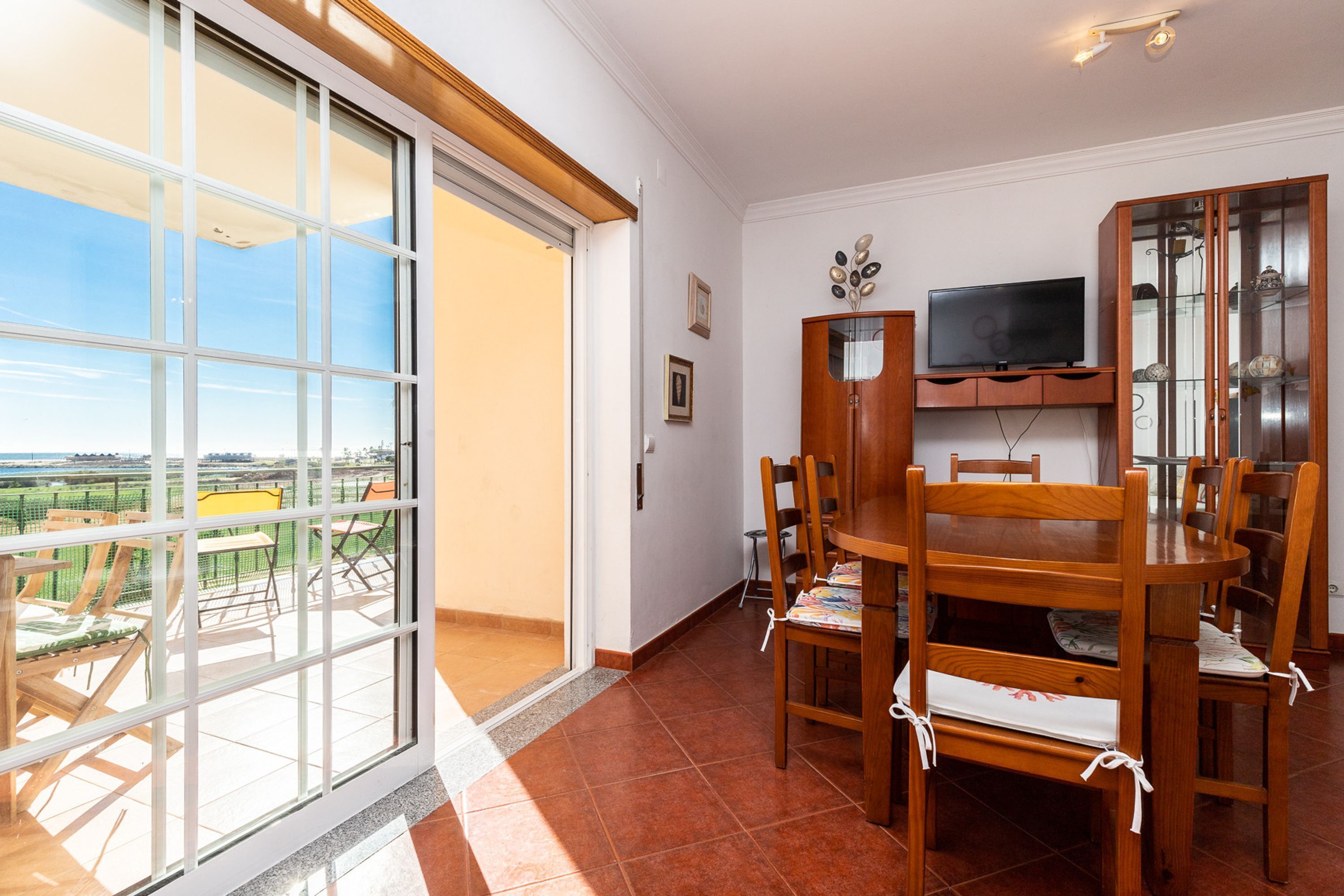 Een eethoek met een grote tafel en stoelen, een aan de muur bevestigde televisie en een schuifdeur naar een balkon met buitenmeubilair.