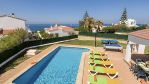Une piscine dotée de chaises longues et d’une table de ping-pong, entourée de verdure et de bâtiments, avec vue sur la mer.