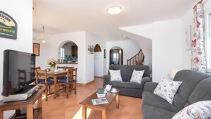 Un vaste espace de vie comprenant un canapé, une table basse et un téléviseur. On y trouve également un coin repas avec des chaises et une table. Un escalier mène à l’étage supérieur.