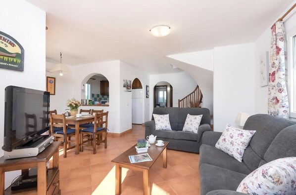A spacious living area with a sofa, coffee table, and TV. There is a dining area with chairs and a table. A staircase leads to an upper level.