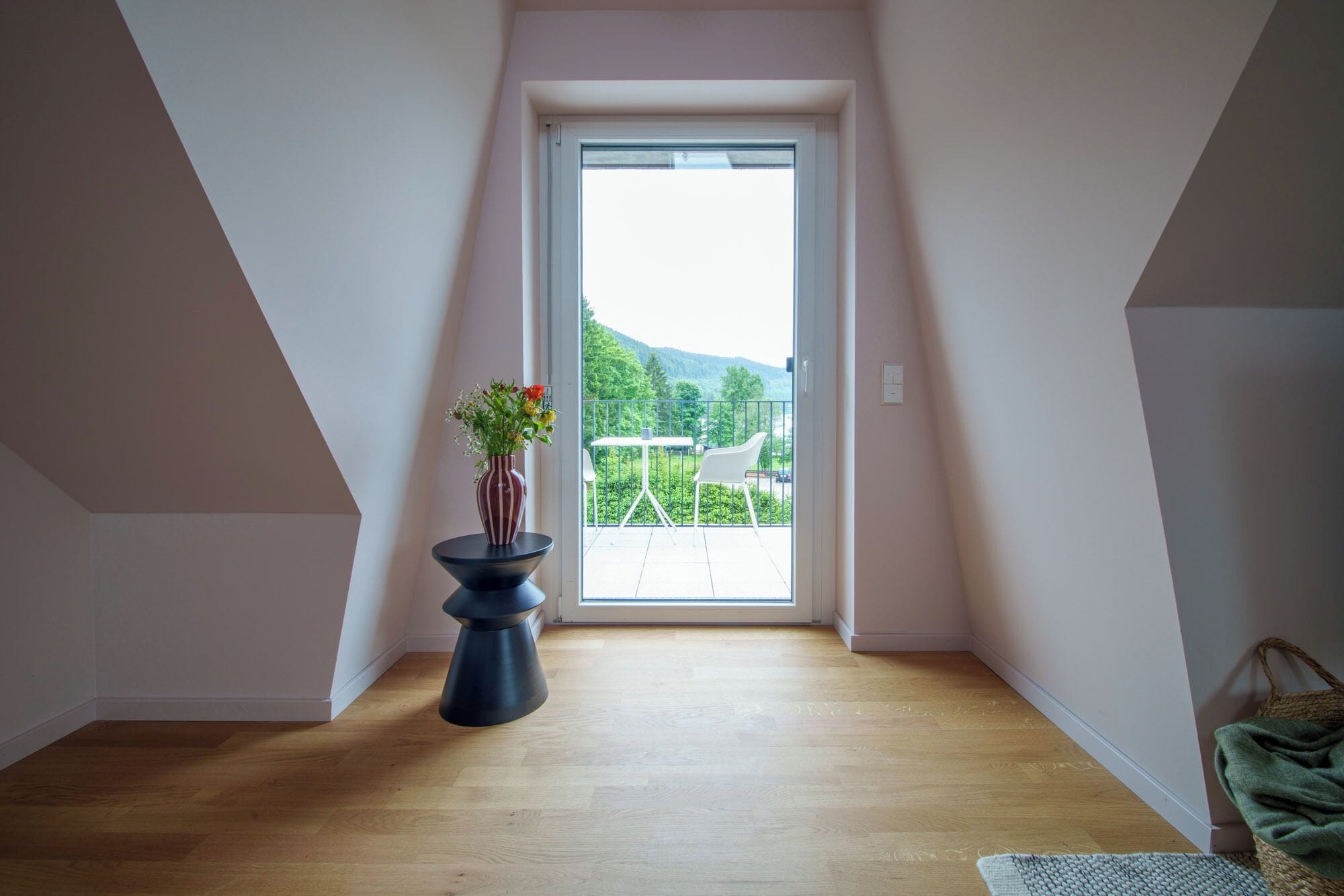 A room with a wooden floor, a vase with flowers, and a view of greenery and mountains through a glass door.