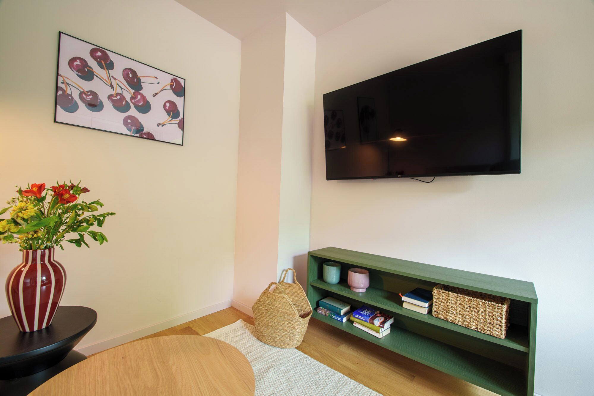 A living room with a flat-screen TV, a wall-mounted picture, a bookshelf, a round wooden table, and a vase with flowers.