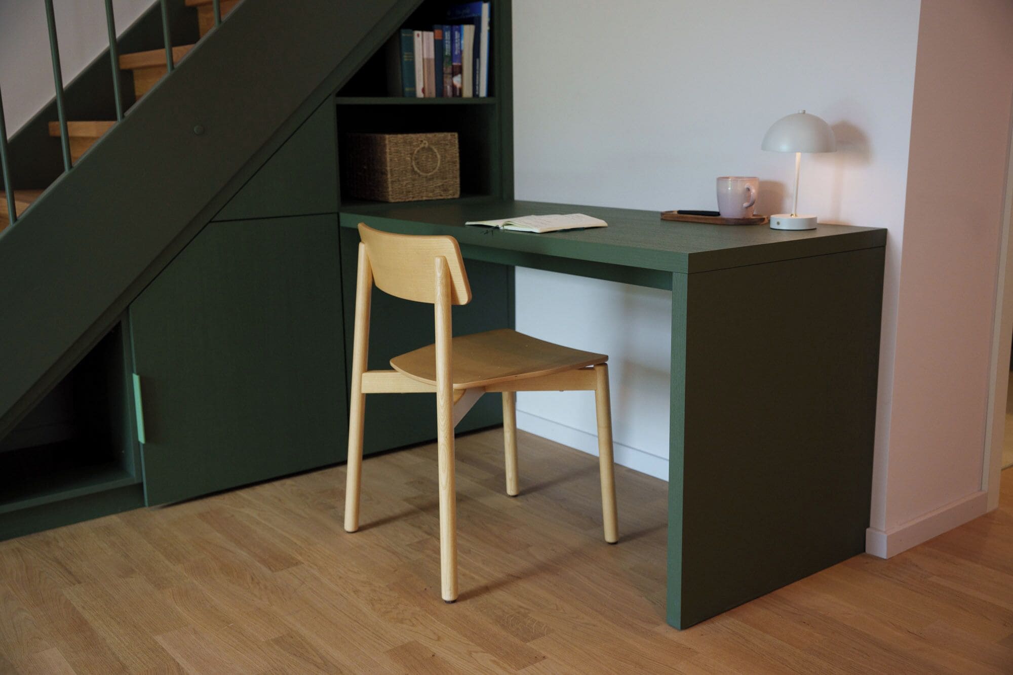 Un petit bureau vert foncé, avec une chaise en bois placée devant. Une lampe se trouve sur le bureau, et un livre est posé sur l’étagère derrière le bureau.
