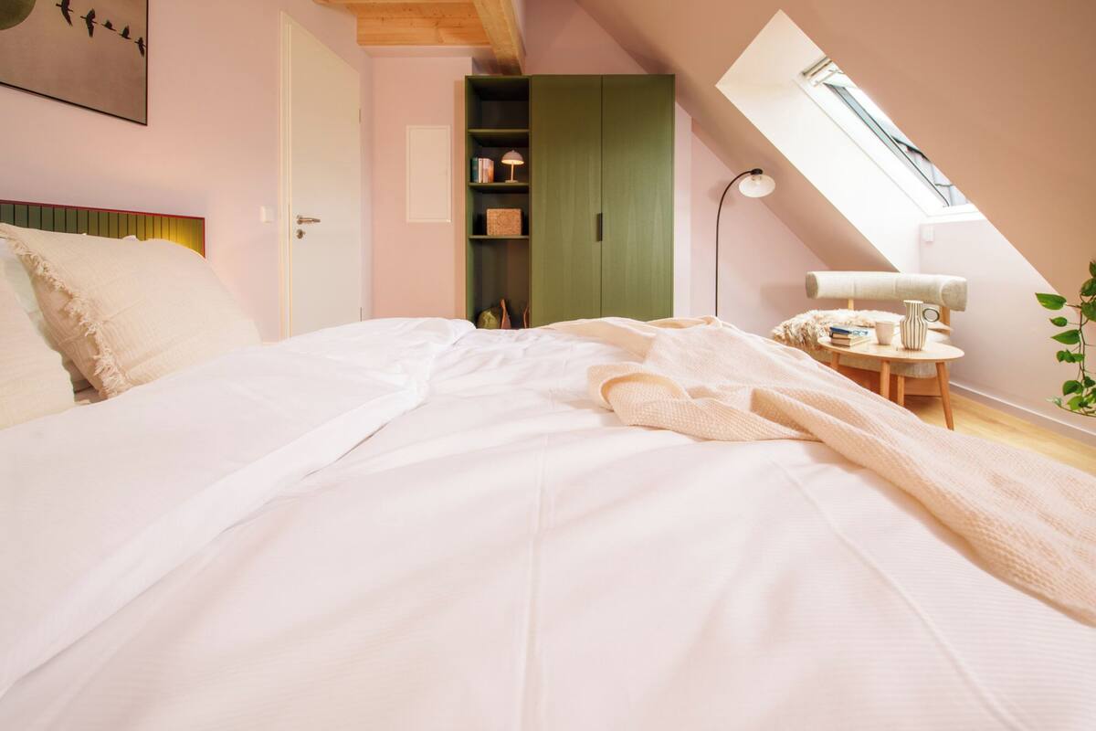 A bedroom with a large bed, a skylight, a green wardrobe, and a small table with a lamp.