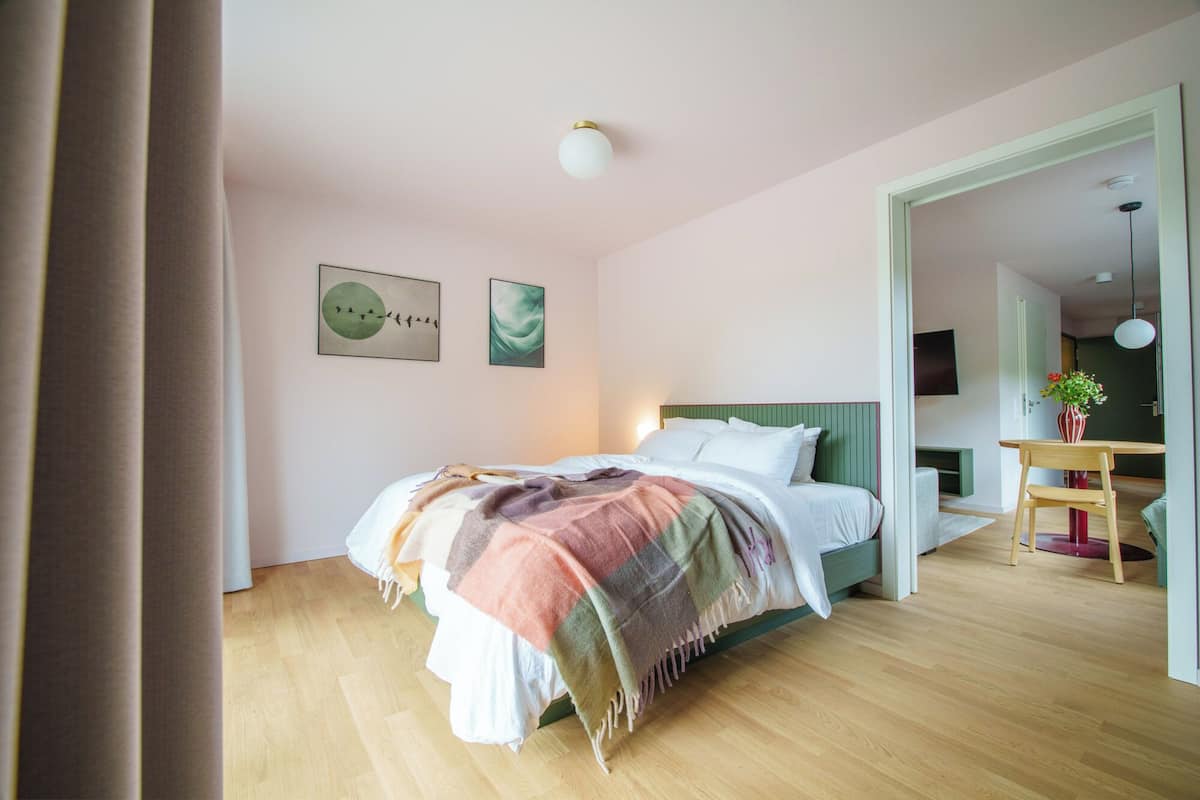 A modern bedroom with a wooden floor, a bed with a colorful blanket, a green headboard, and two wall art pieces.