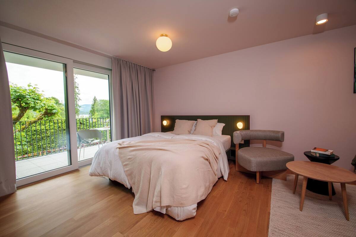 A bedroom with a large bed, a chair, a small table, and a view of greenery through a sliding door.
