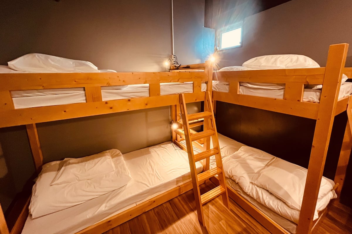 Une chambre avec un lit superposé, une échelle, un plancher en bois et une fenêtre laissant entrer la lumière naturelle.